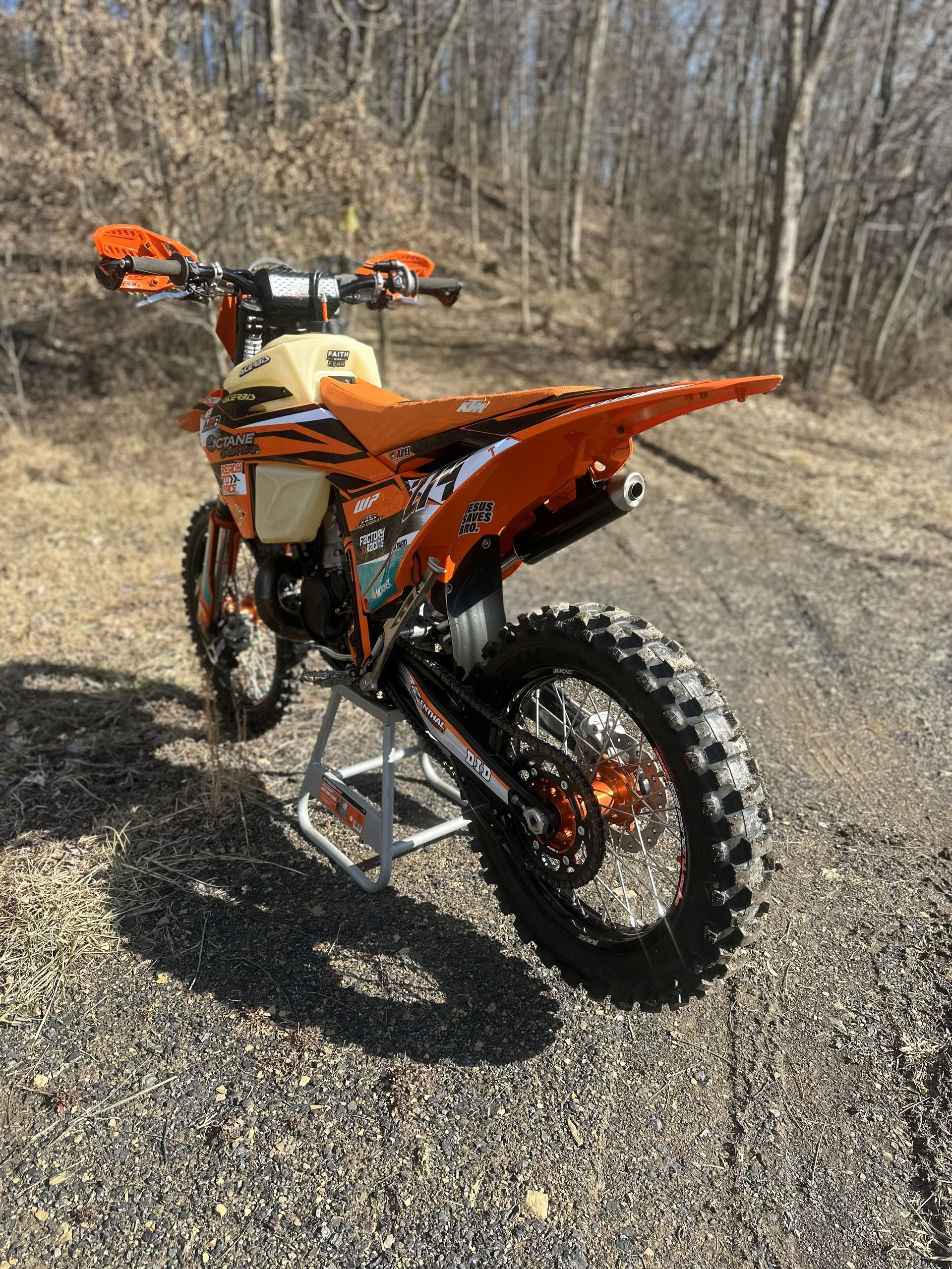 Orange and black dirt bike on a stand outdoors with a dirt trail in the background.