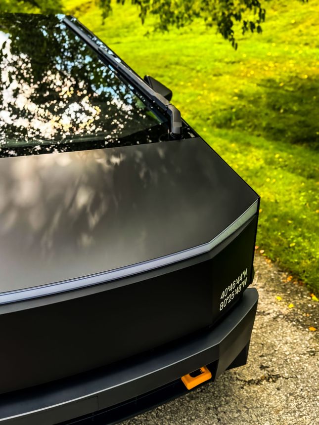 Matte black Tesla Cybertruck with orange tow hook, parked on a path with green grass.