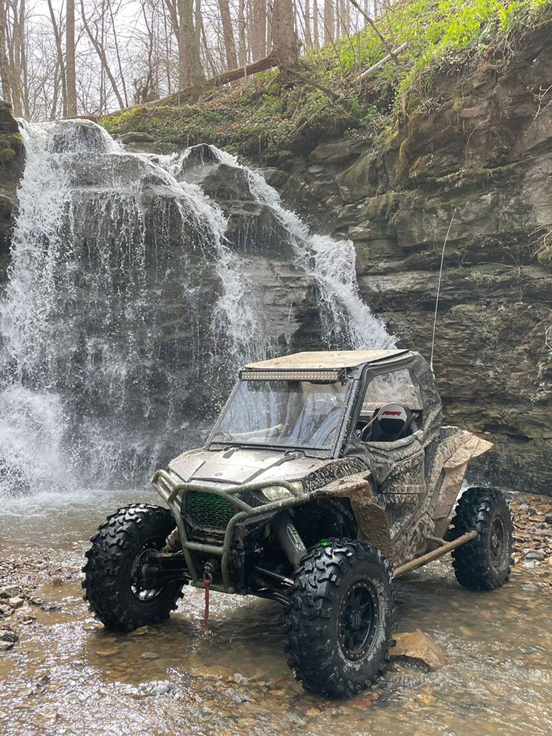 Muddy UTV parked in shallow water near a small waterfall in a forest.