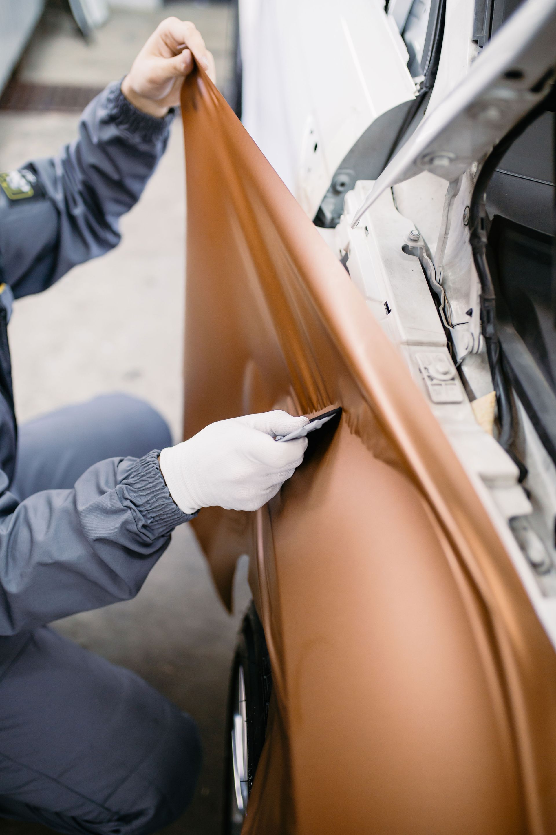 Person in gray jumpsuit applying bronze vinyl wrap to a car door with a blade.