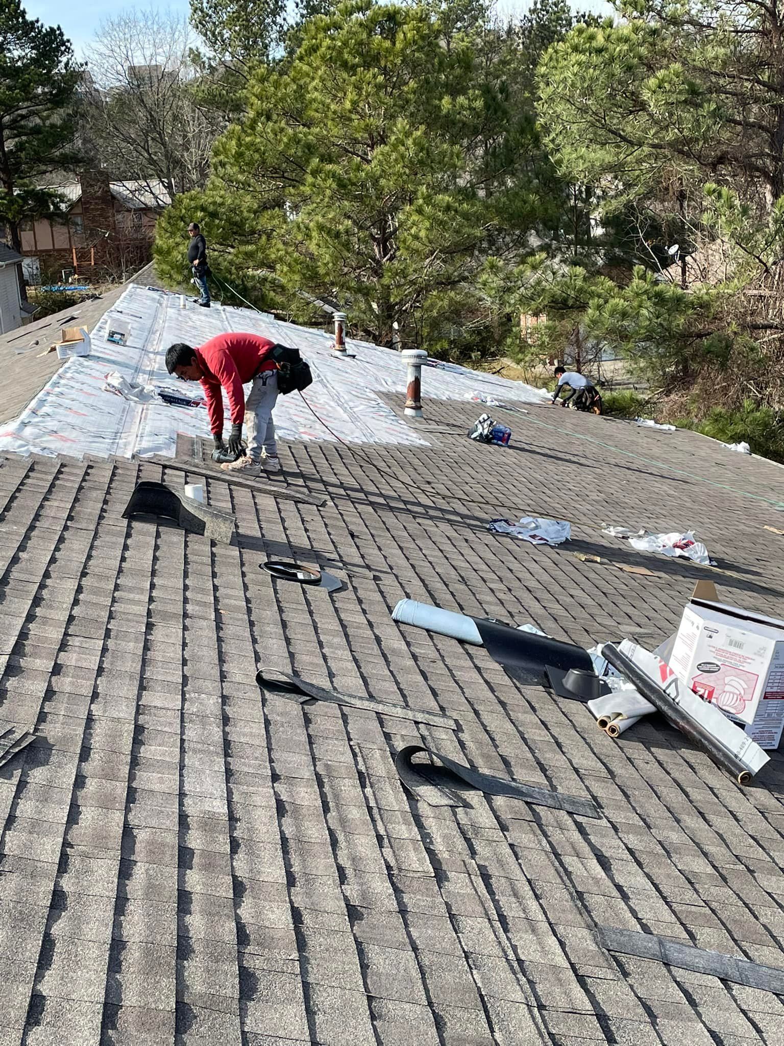 Roofers working on a shingled roof, trees in the background. Bright day, red shirt.