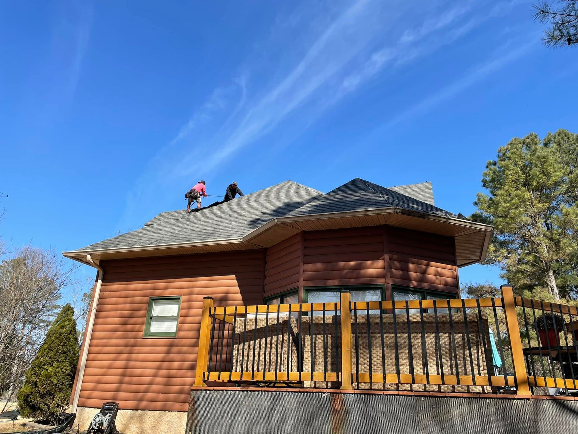 Two people on a brown house roof, working. A blue sky and wooden deck are visible.