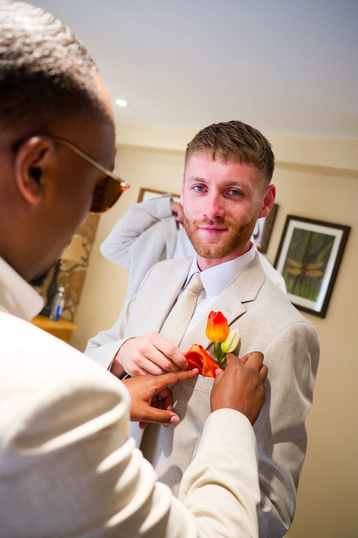 A person adjusts a bright orange and yellow floral boutonnière on the lapel of a light-colored suit jacket.
