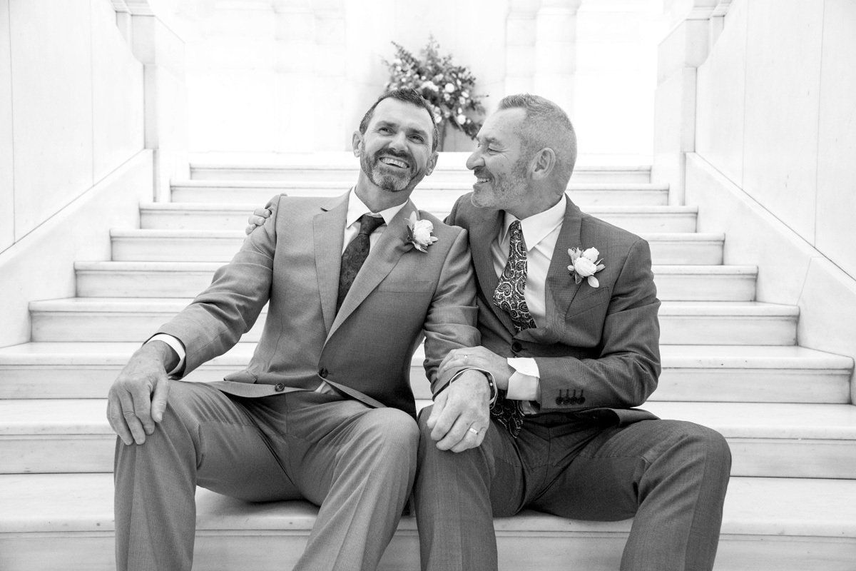Two men in suits are sitting on a set of stairs holding hands.