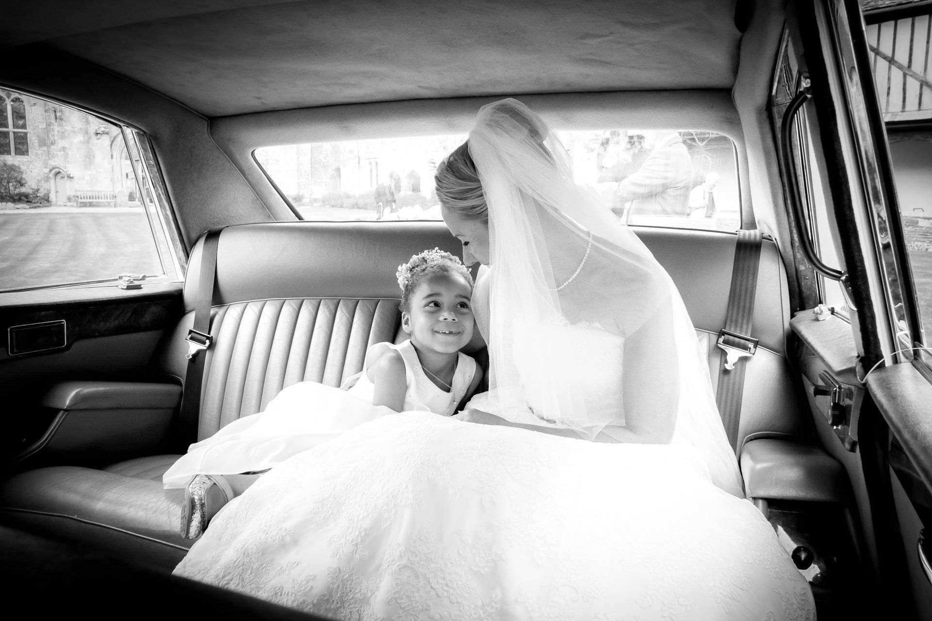 A bride and a flower girl are sitting in the back seat of a car.