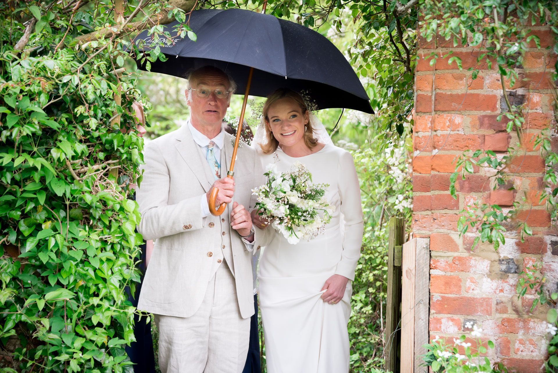 A bride and groom are standing under an umbrella in the rain.