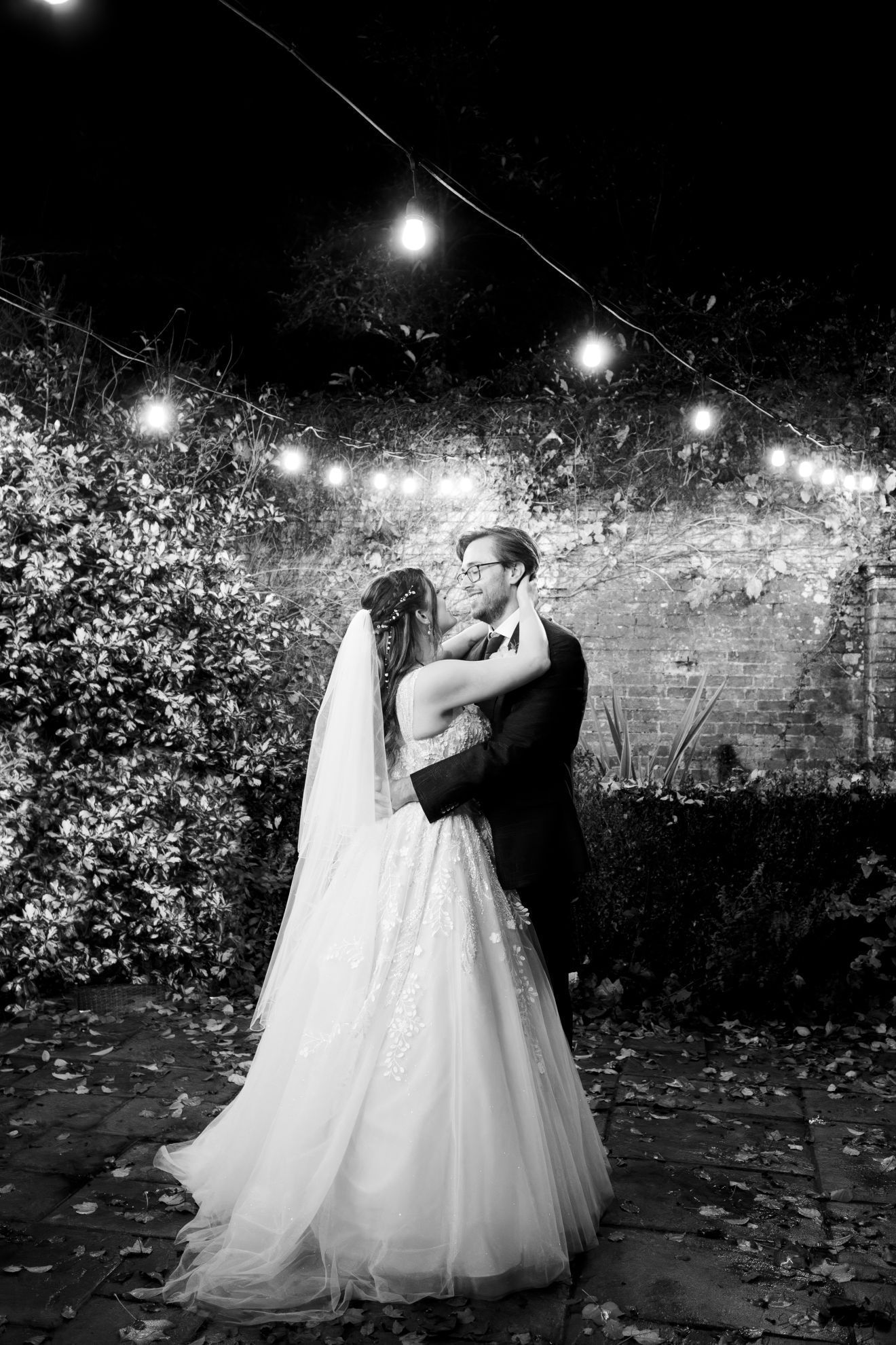 A black and white photo of a bride and groom dancing under a string of lights.