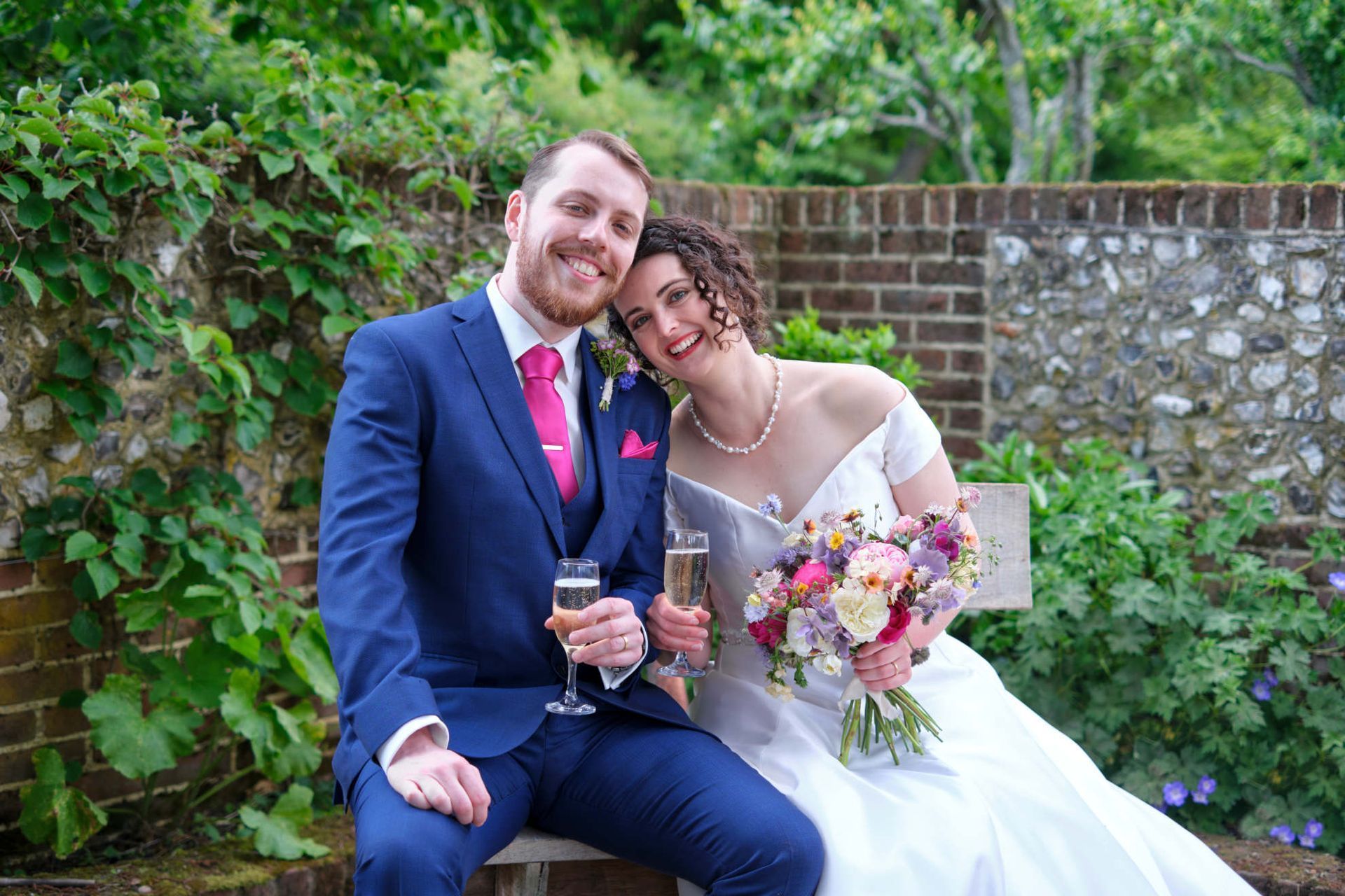 A bride and groom are sitting on a bench holding champagne glasses.