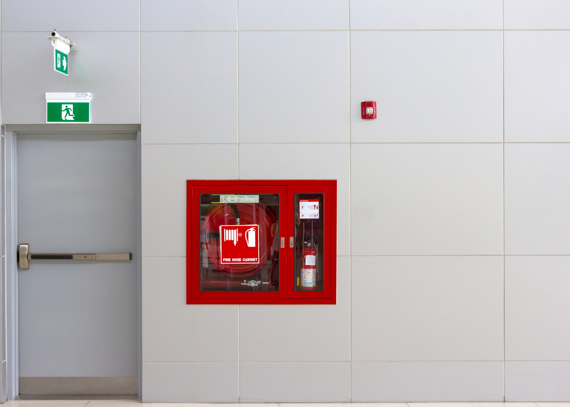Gray door with exit sign above, next to a fire extinguisher and alarm on a white wall.