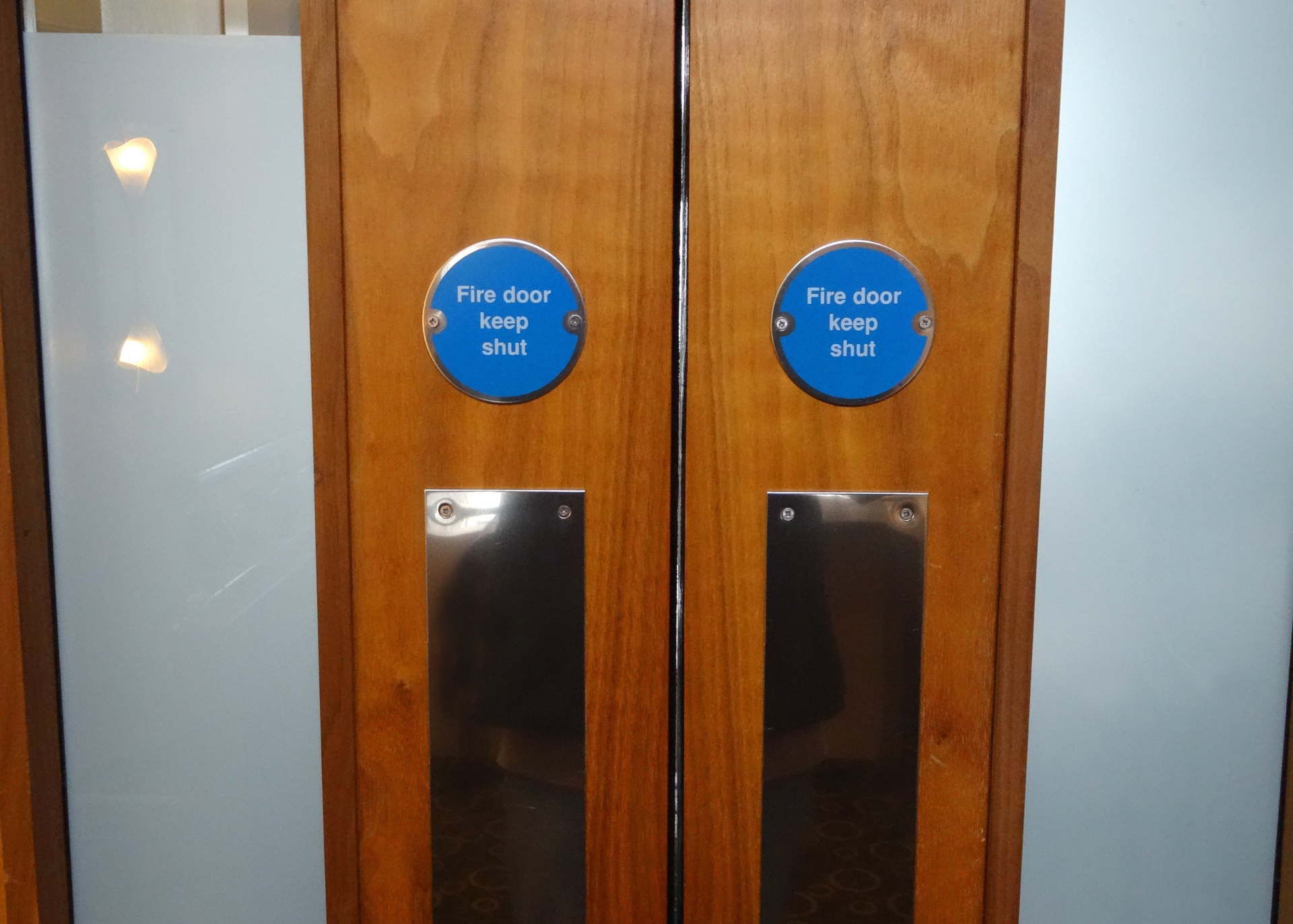 Wooden double doors with frosted glass panels, blue fire door signs, and metal handles.