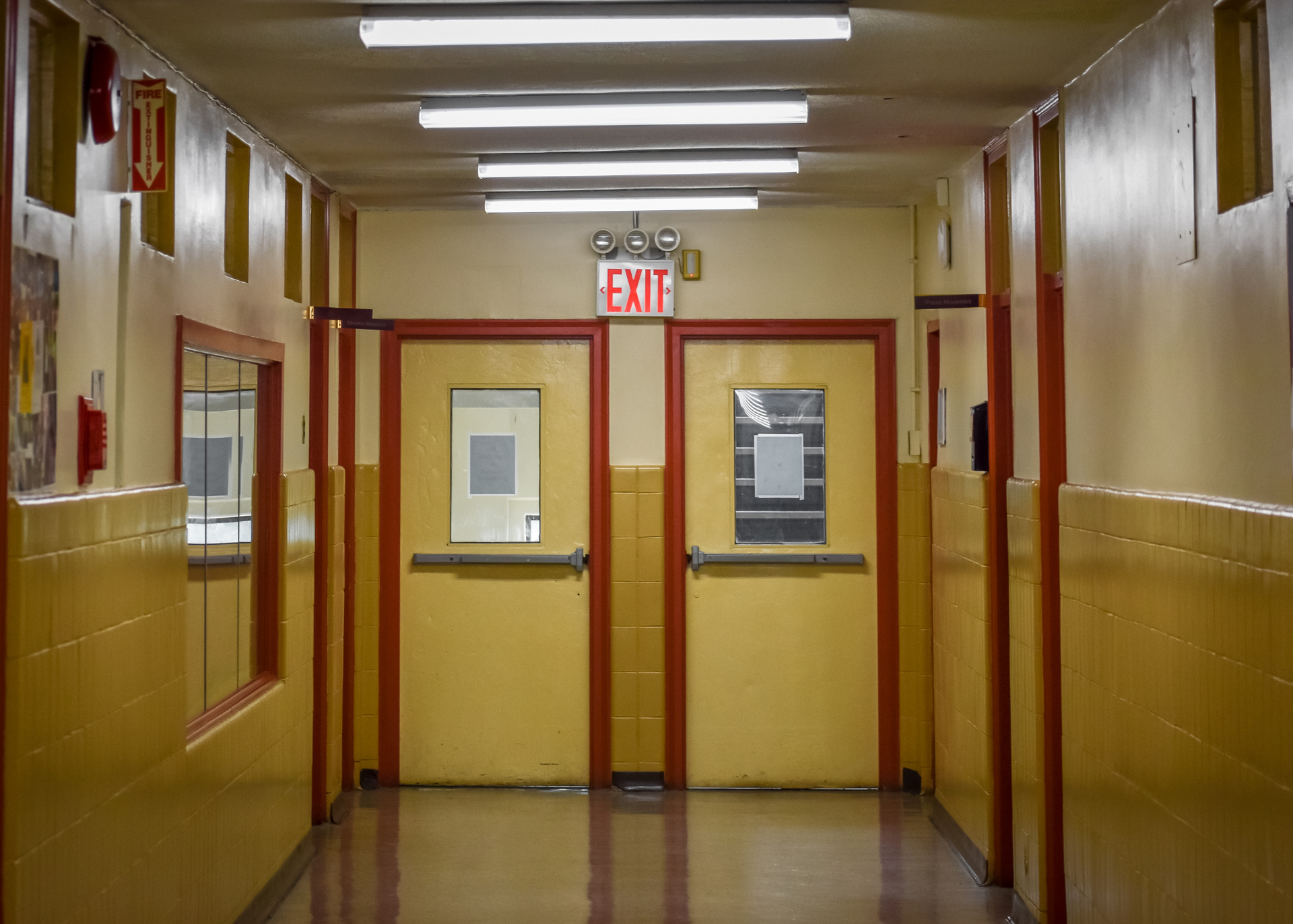School hallway with yellow walls, red trim, closed yellow doors, and an exit sign.