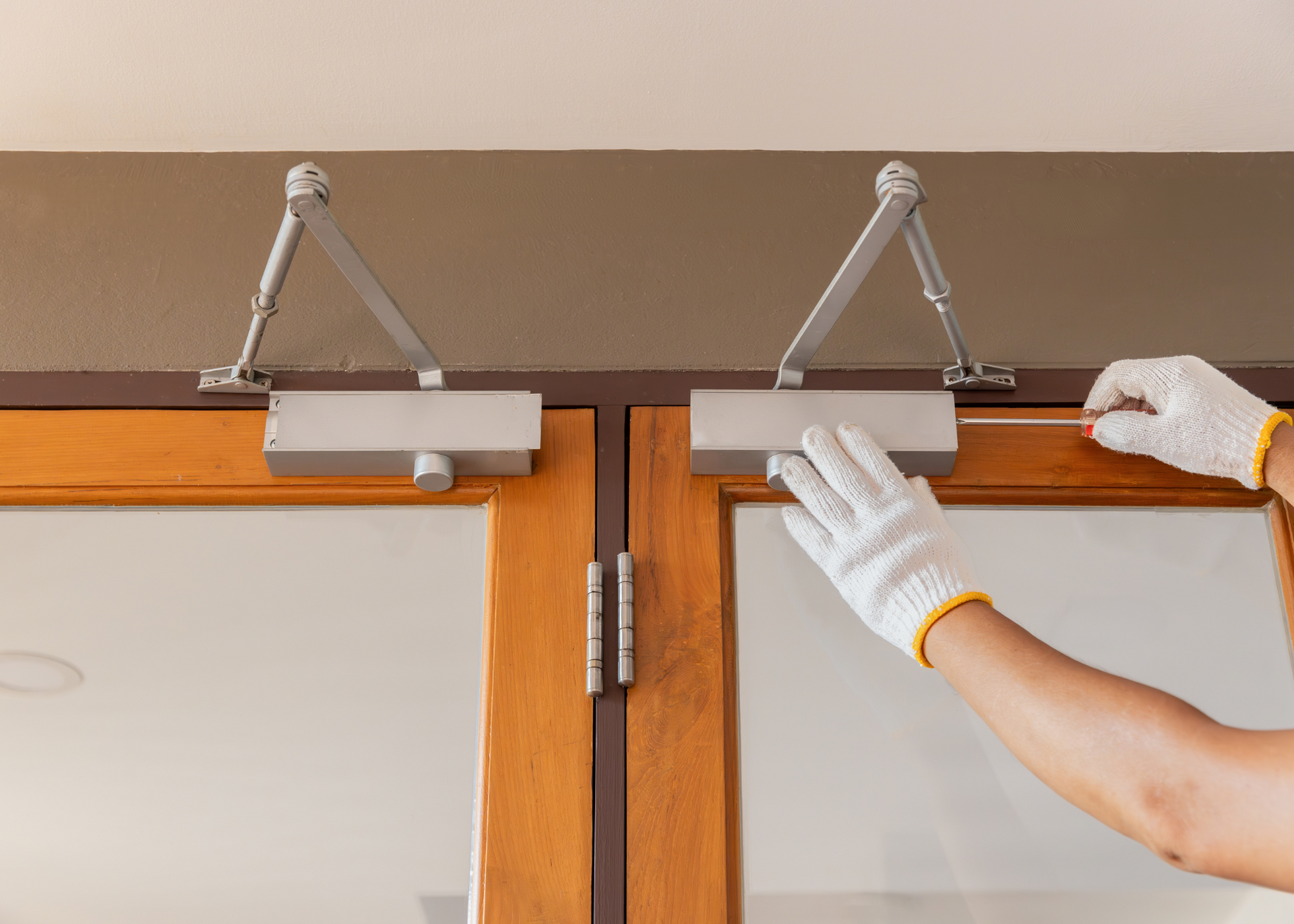 Person adjusting a door closer on a wooden door with white gloves, indoors.