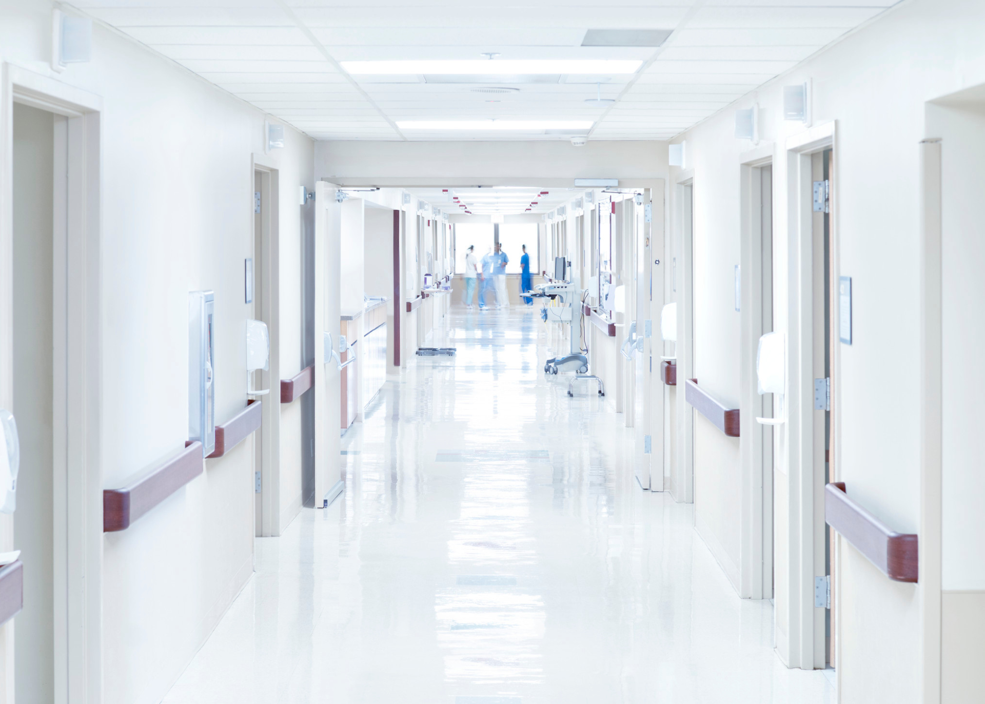 Bright empty hospital hallway with white walls, open doors, and a person in blue in the distance