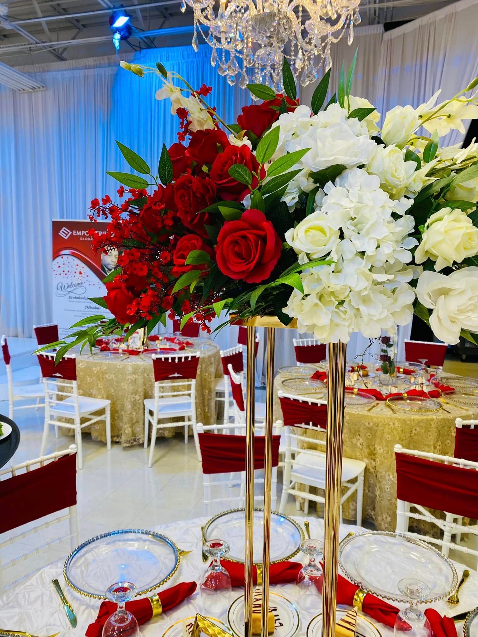 A banquet room features gold-clothed tables, white chairs with red covers, and a large red and white floral centerpiece.