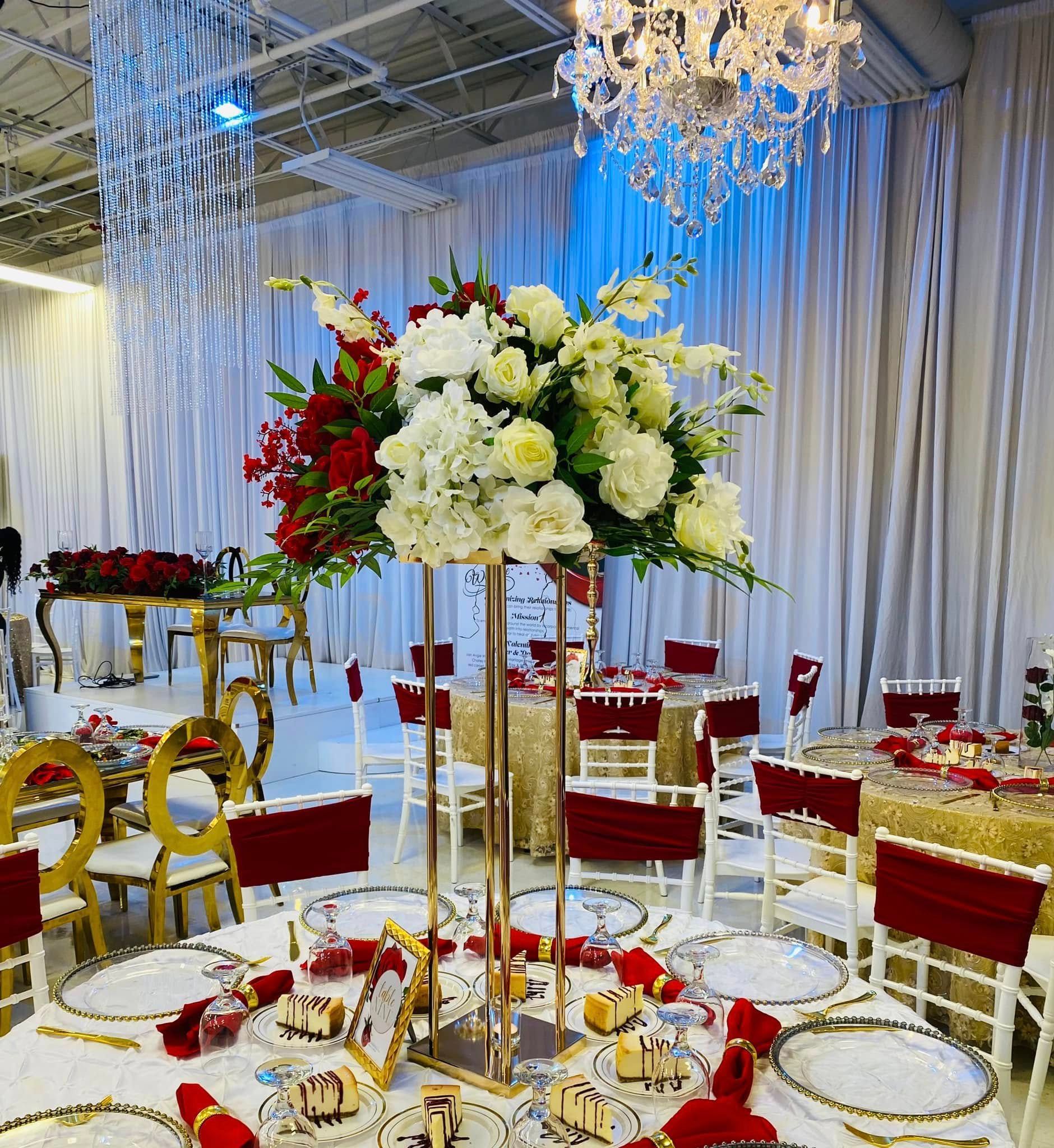 A formal banquet hall with round tables, gold chairs, red accents, and a large white floral centerpiece on a tall stand.