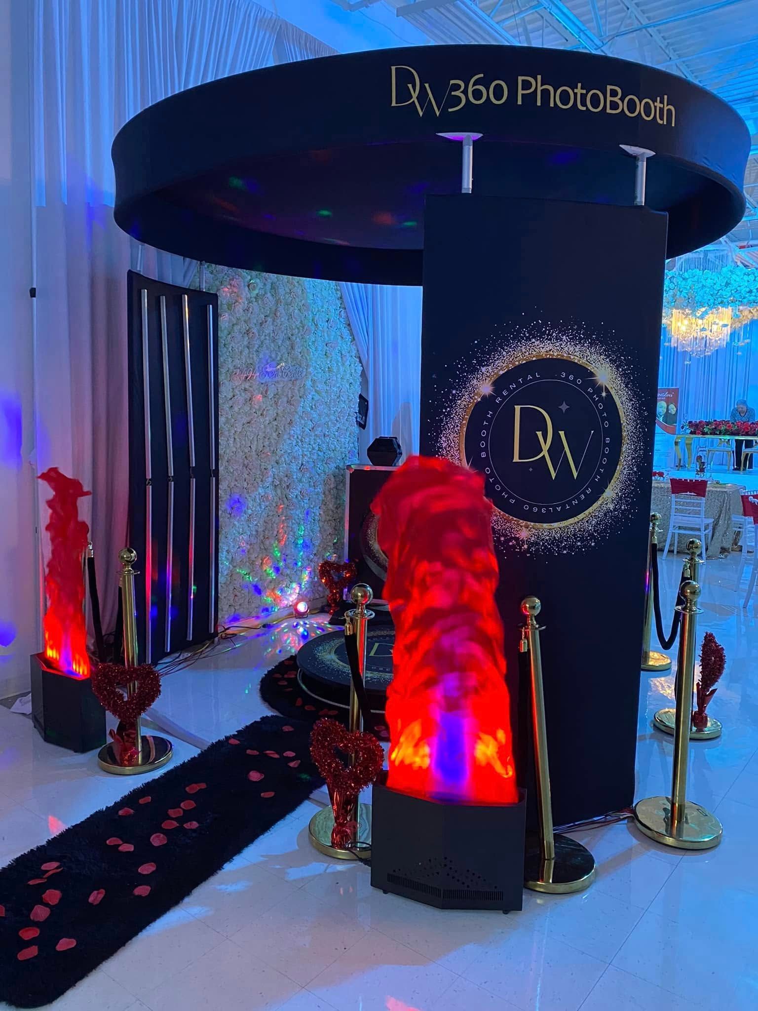 A 360 photo booth setup with a circular overhead sign, a branded backdrop, red flame lights, and a velvet red carpet.
