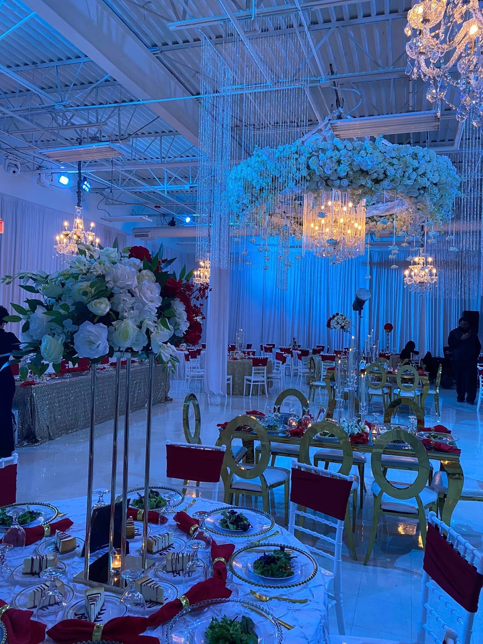 A grand wedding reception hall with tables featuring floral centerpieces, crystal chandeliers, and red accent chairs.