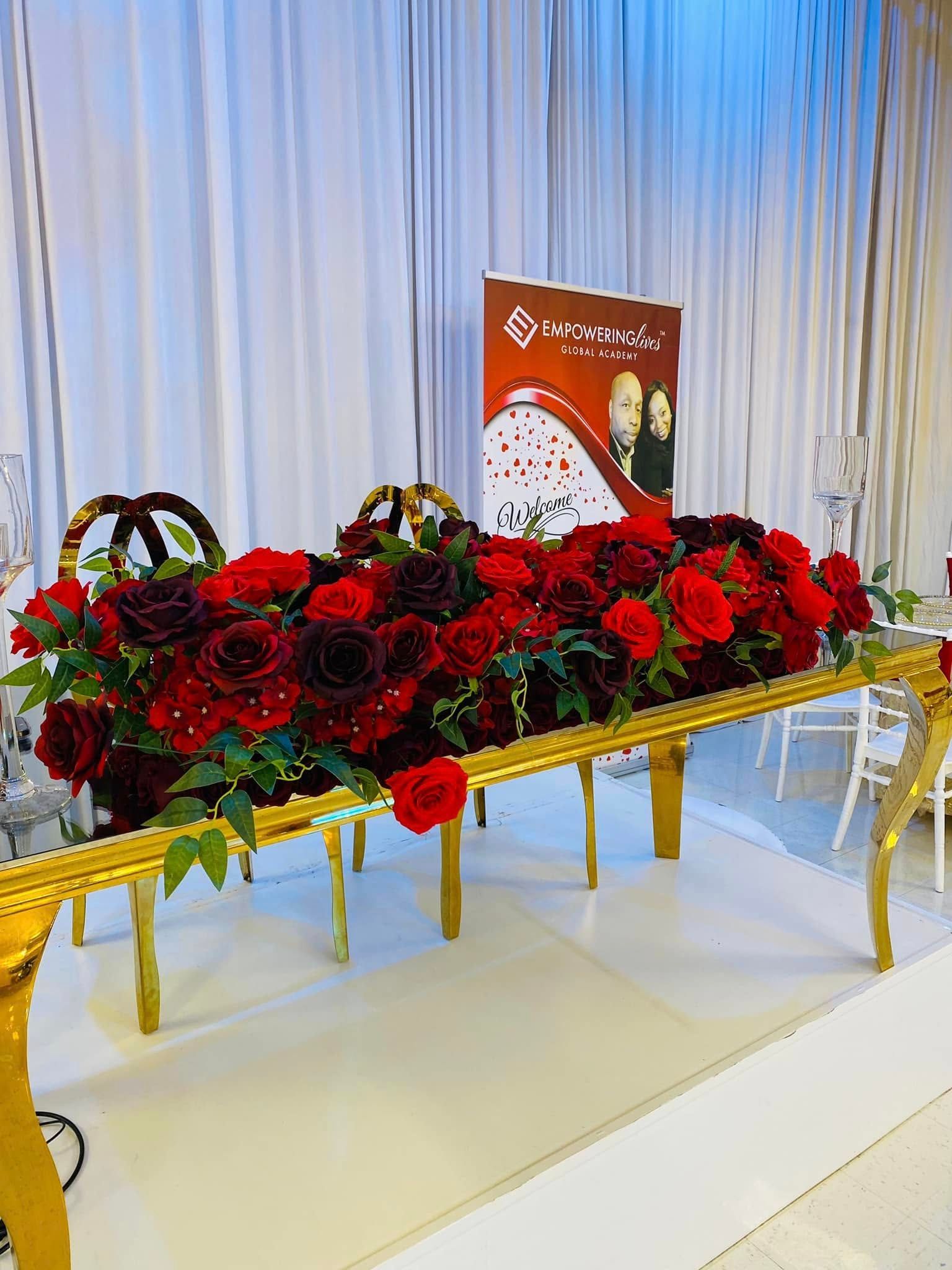 A table decorated with vibrant red roses, gold accents, and a brand banner, set before white curtains.