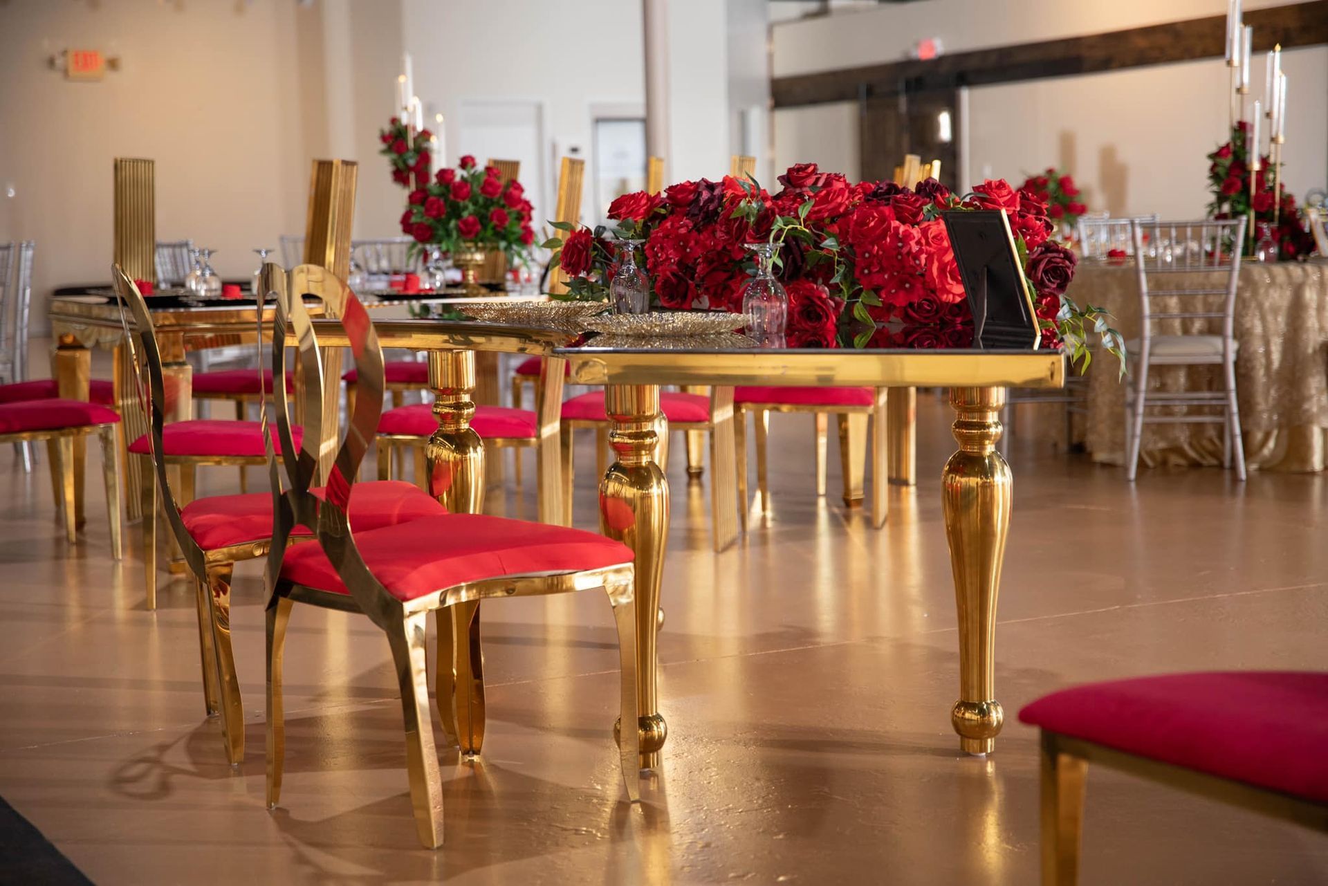Gold-framed chairs with red velvet seats surround a long table set with vibrant red floral arrangements in a banquet hall.