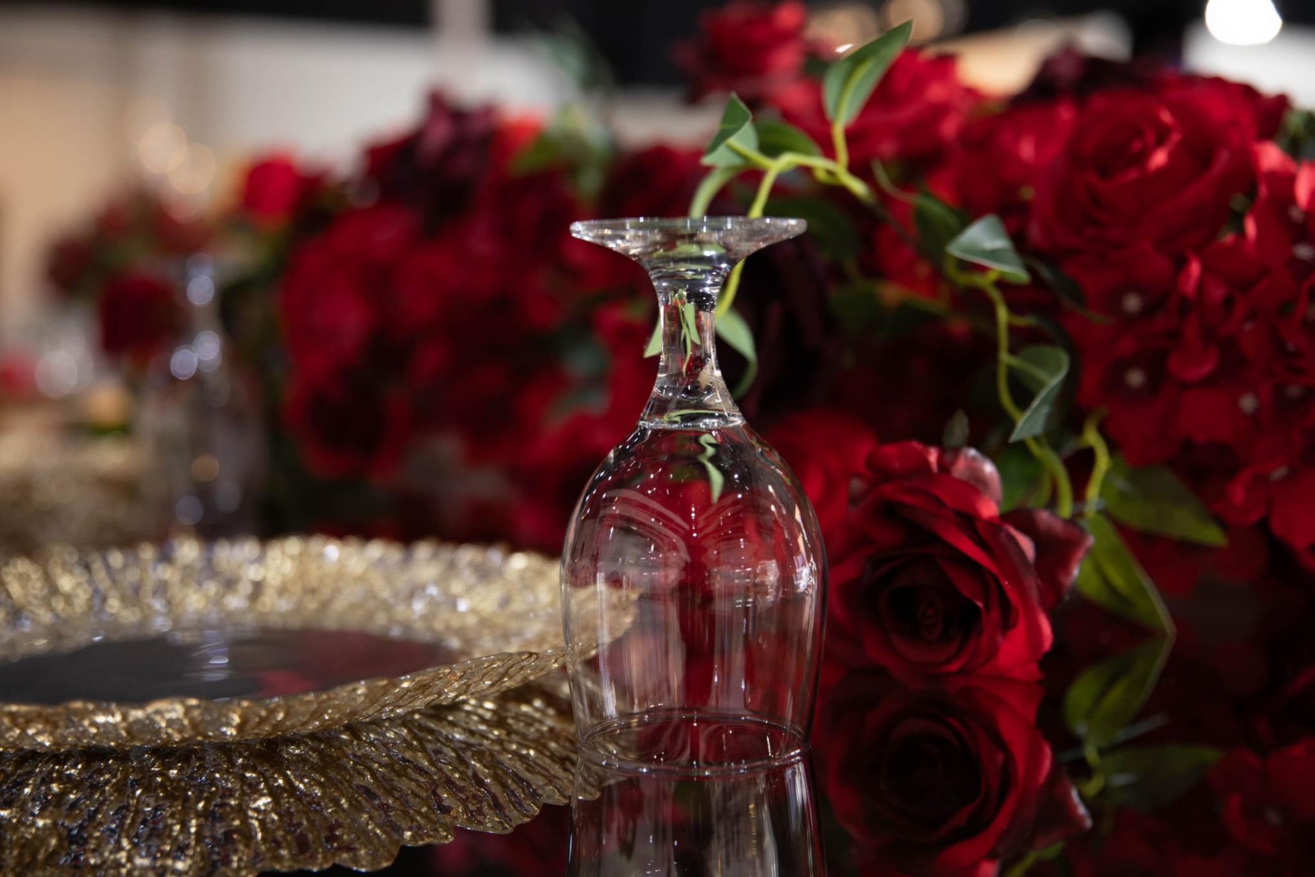 A clear glass goblet upside down on a table next to a gold textured charger plate, surrounded by deep red roses.