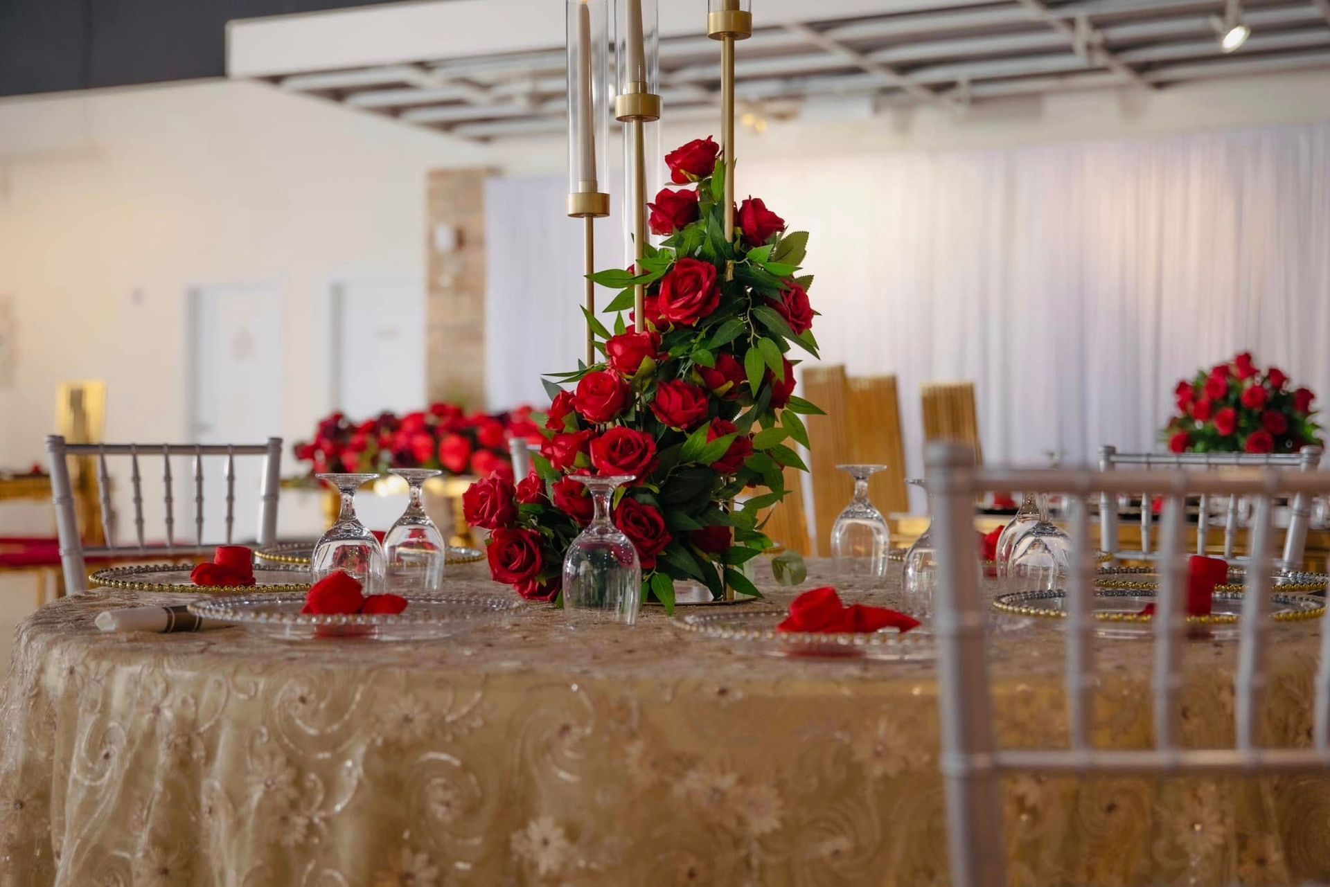 A round banquet table with a gold textured cloth, decorated with red roses and a tall centerpiece in a bright event space.