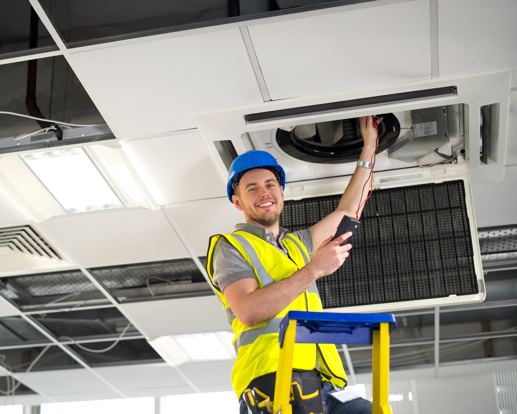 HVAC technician in hard hat, safety vest, on ladder, inspecting ceiling unit, smiling.