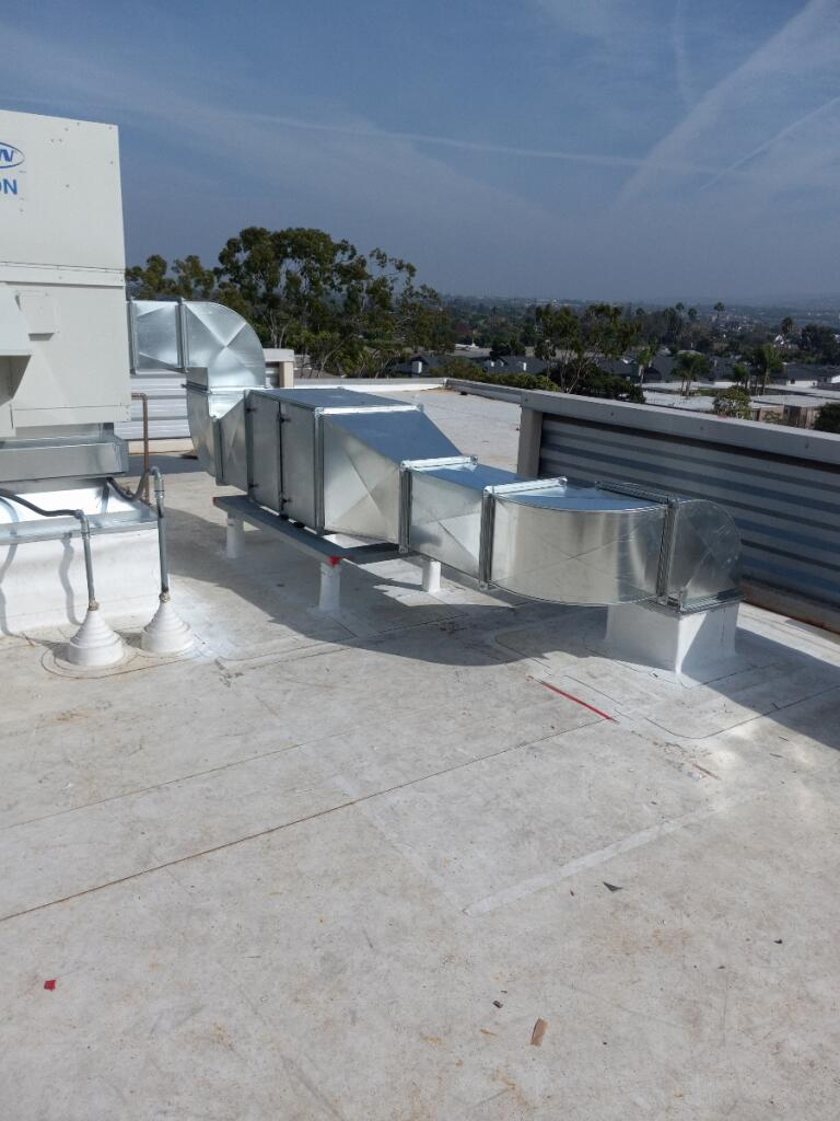 Rooftop with HVAC ductwork, white and silver against a blue sky with a distant tree line and city view.