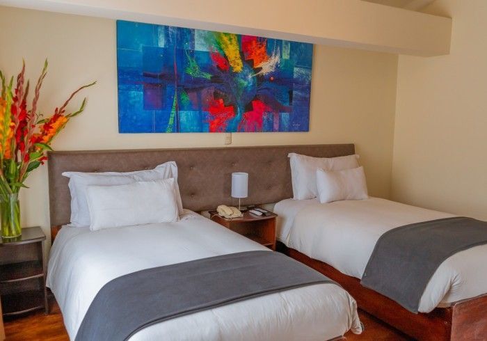Habitación de hotel con dos camas individuales, sábanas blancas, mantas grises y coloridos cuadros en la pared