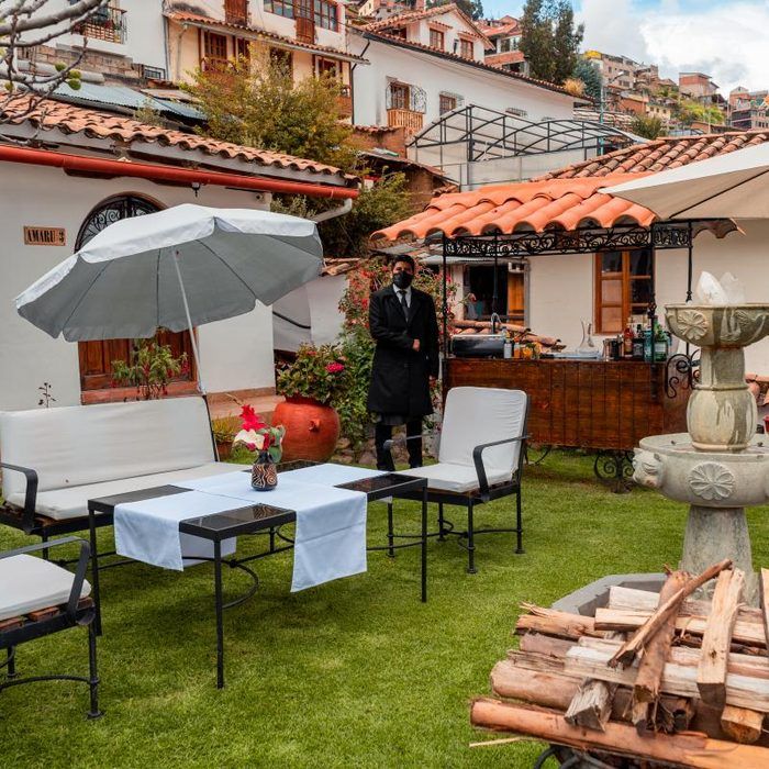 Patio exterior con mesas, sillas, sombrilla y fogata en un patio cubierto de césped cerca de edificios con techos de