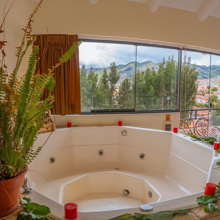 Bañera de hidromasaje blanca junto a una ventana con vistas a la montaña, velas rojas y una planta en maceta.