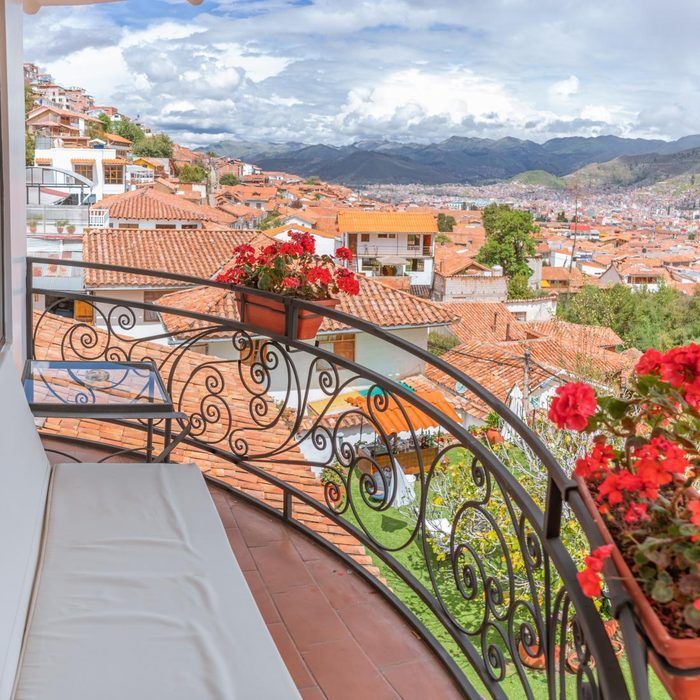 Balcón con silla blanca, flores rojas y vista de tejados de terracota y montañas bajo un cielo nublado.