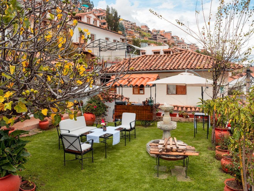 Acogedor patio ajardinado con sillas blancas, mesitas, macetas con flores y una casa con tejado de tejas rojas al fondo.