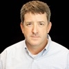 A man in a blue shirt is standing in front of a black background.