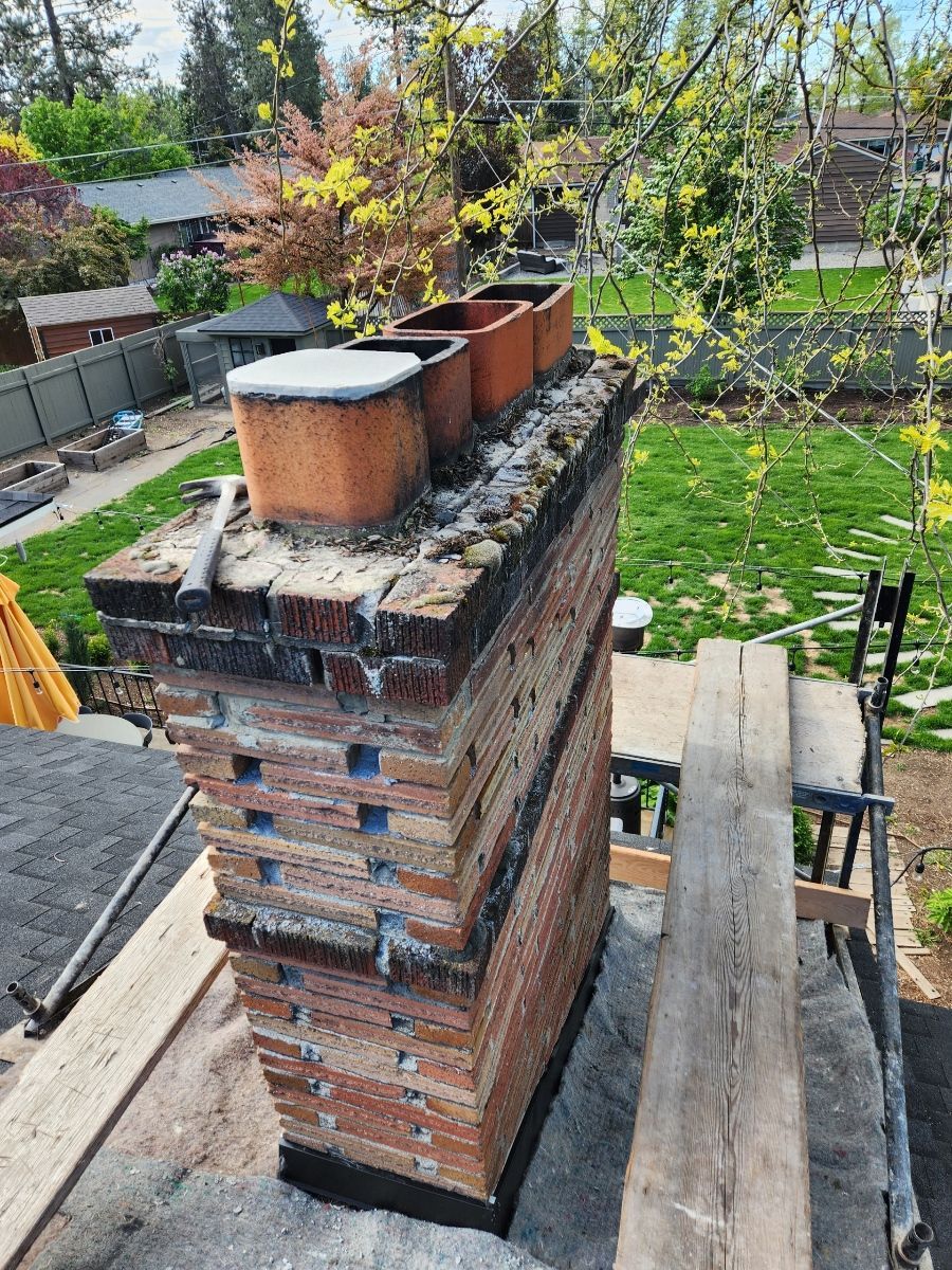 Brick chimney on a roof with a damaged top and three flue caps. Green yard in the background.