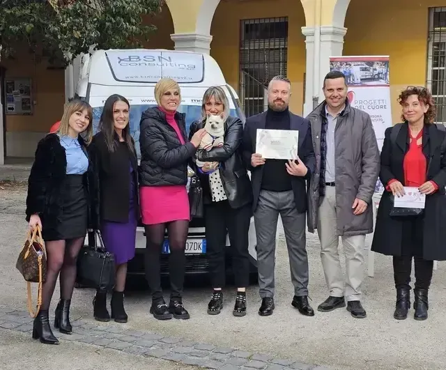 Sette persone in piedi davanti a un edificio, una con in mano un certificato e un'altra con un cagnolino.
