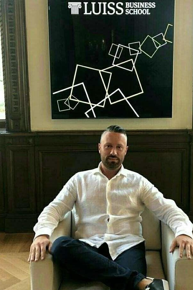 Un uomo seduto in poltrona sotto l'insegna della LUISS Business School in una stanza scarsamente illuminata.