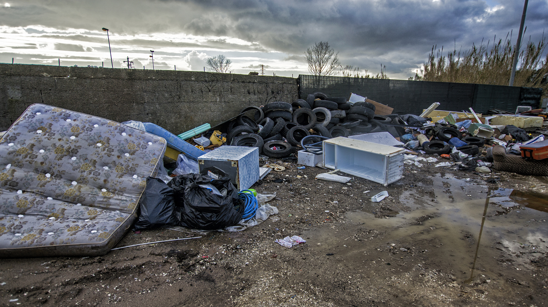 Materassi, pneumatici e detriti abbandonati ammucchiati in una discarica fangosa sotto un cielo nuvoloso.