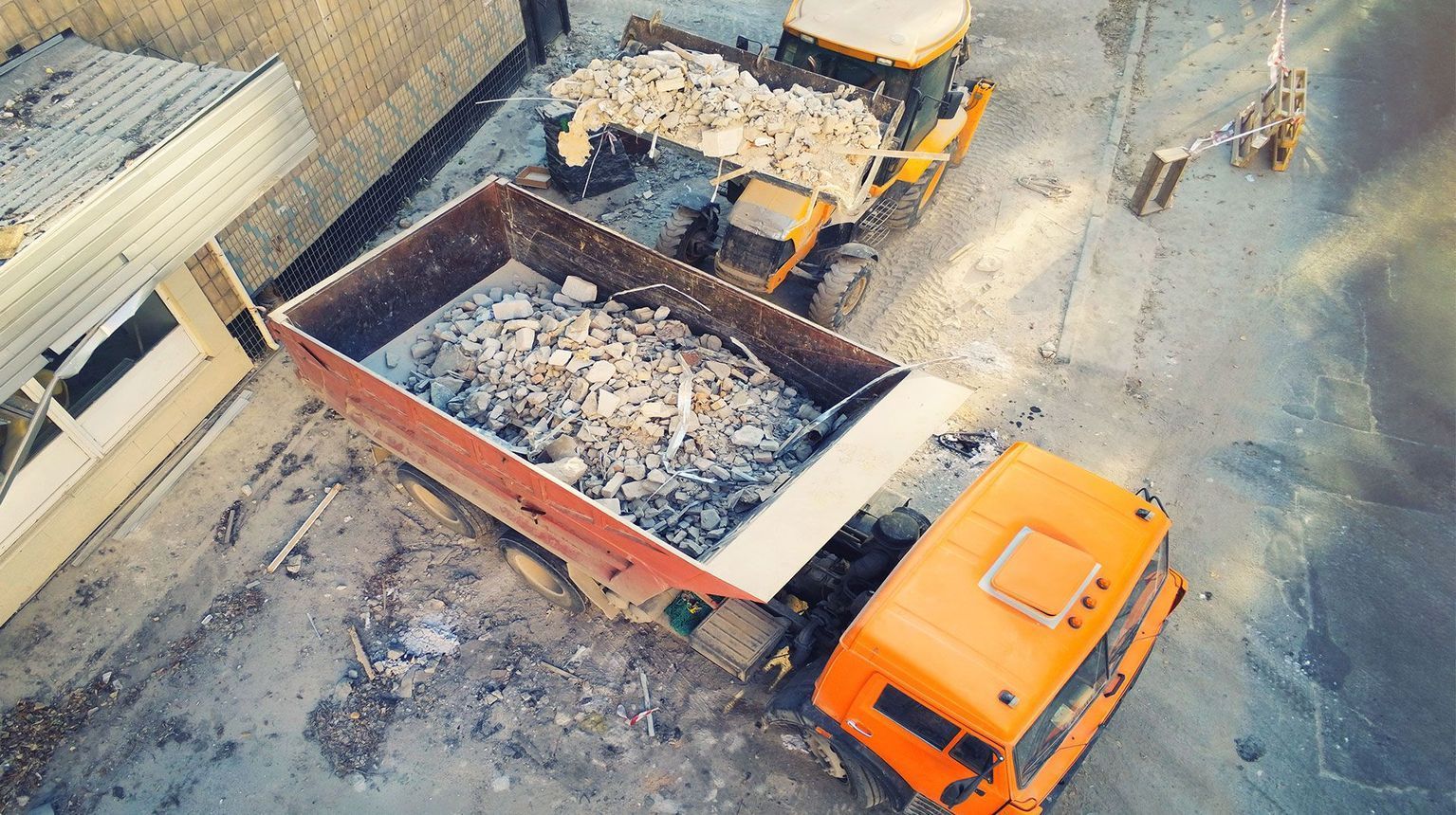 Un camion ribaltabile arancione carico di macerie in un cantiere, visto dall'alto.