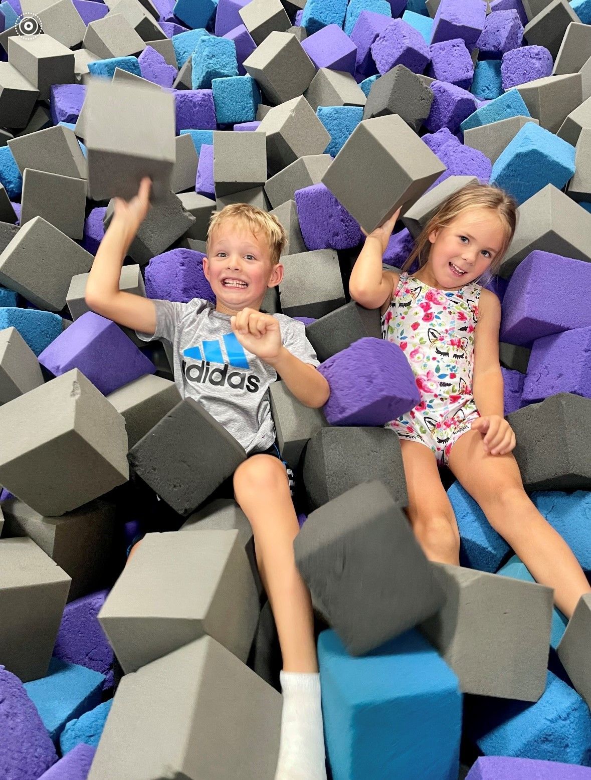 Two children smiling in a foam pit, surrounded by gray, purple, and blue foam blocks