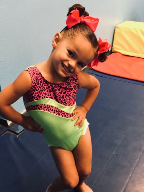 Young child in a pink and green leotard posing with a red bow in a gymnasium