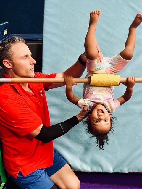 Coach in red shirt holding a child upside down on a balance beam in a gymnasium