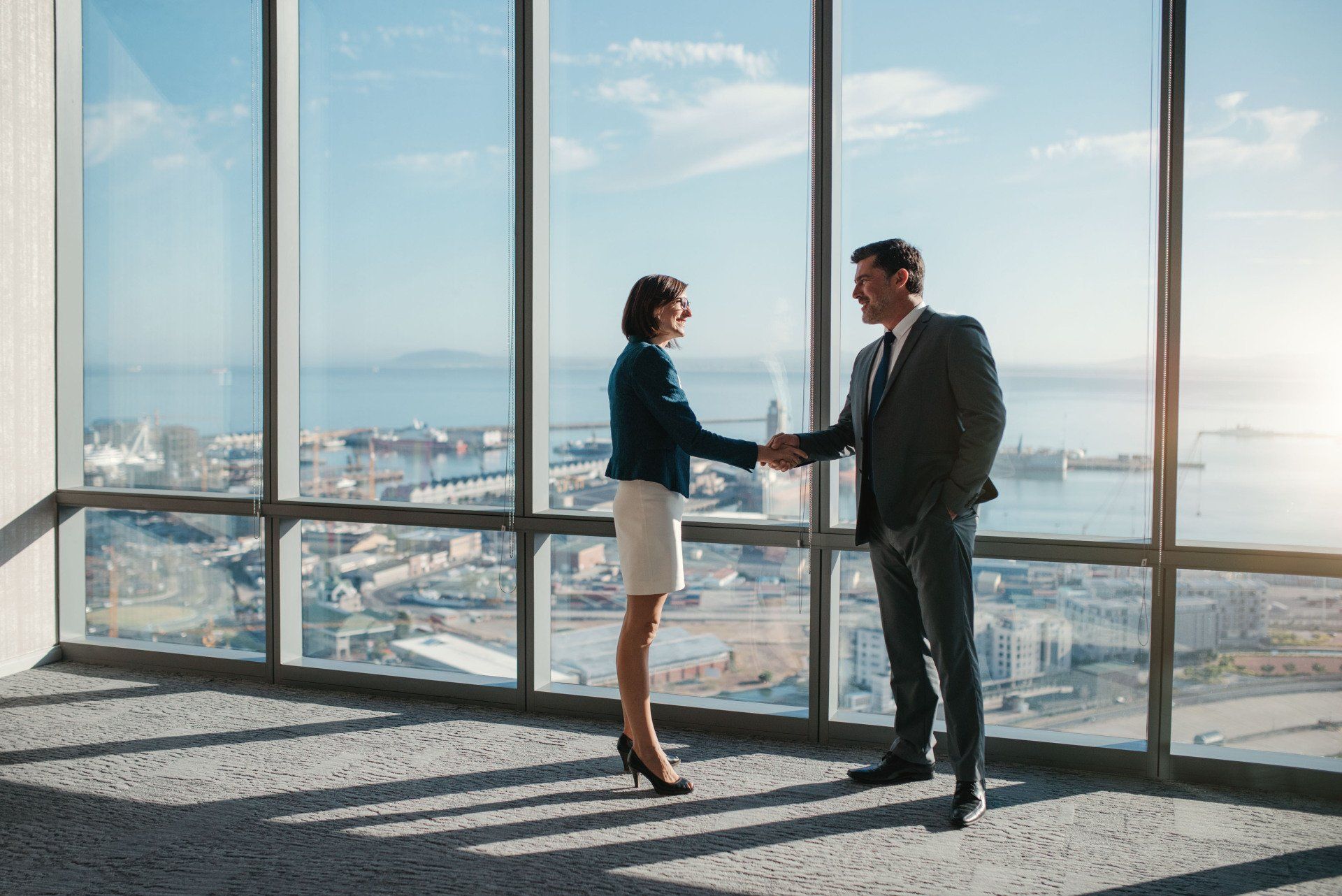 A man and a woman are shaking hands in front of a large window.