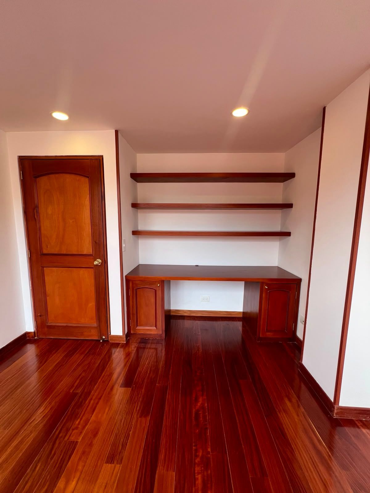 Habitación vacía con suelo de madera pulida, rincón de escritorio, estanterías y una puerta de madera a la izquierda.