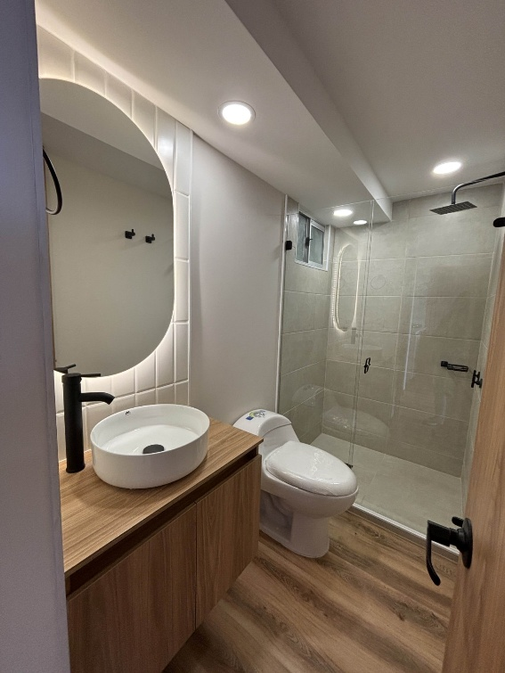 Baño moderno con tocador de madera, lavabo redondo, inodoro y cabina de ducha de cristal.