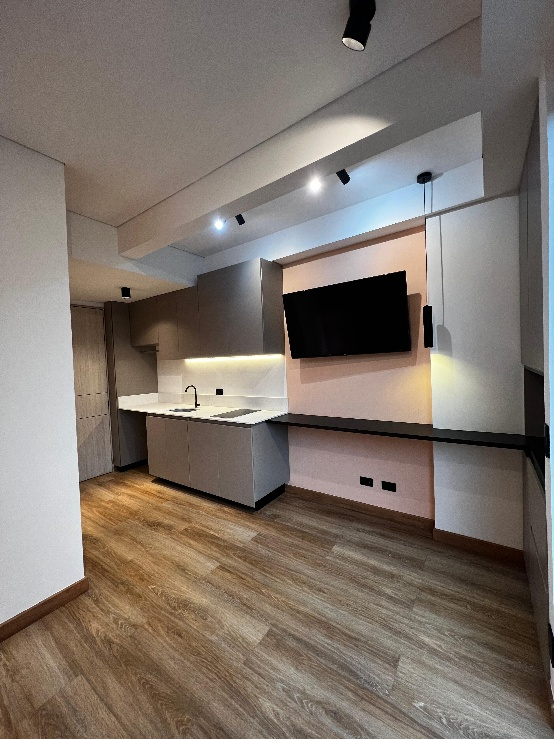 Cocina moderna con suelo de madera, armarios blancos, encimera negra y televisor montado en la pared principal.