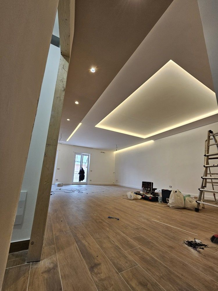 Interior moderno en construcción con suelo de madera, lámparas de techo geométricas, escalera y herramientas a la derecha.