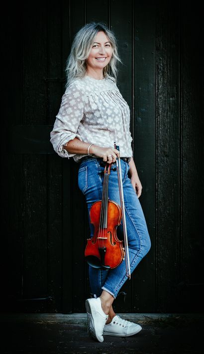 Woman with blonde hair holding a violin, smiling, against a dark wooden door. Wearing jeans, shirt, and sneakers.