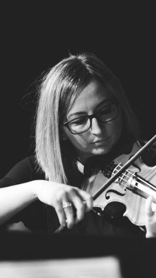 Woman playing violin, wearing glasses, focused expression, in a dark setting.