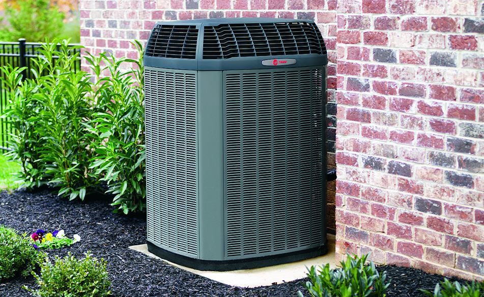 Outdoor air conditioning unit next to a brick wall and shrubbery.