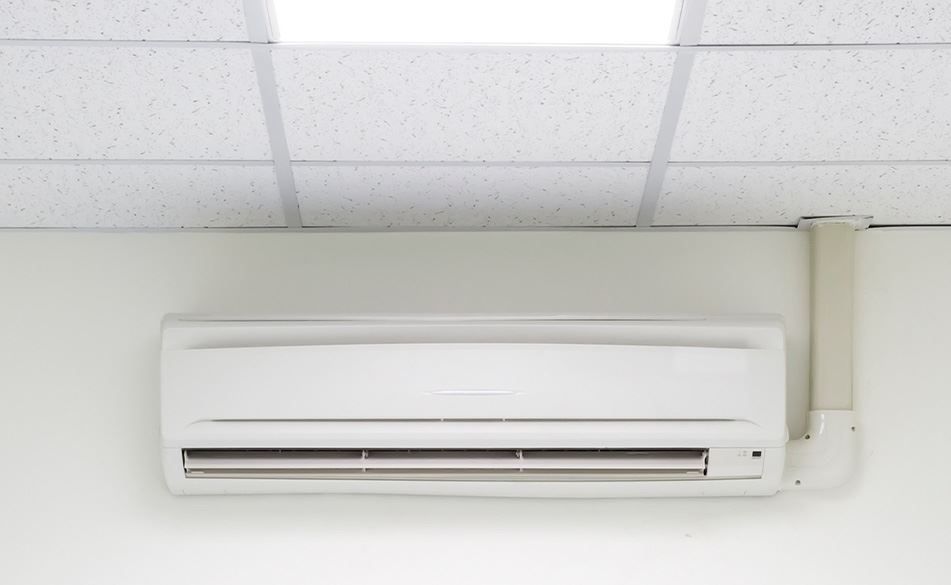 White wall-mounted air conditioner unit with a tube extending to the ceiling, beneath a white ceiling with panels.